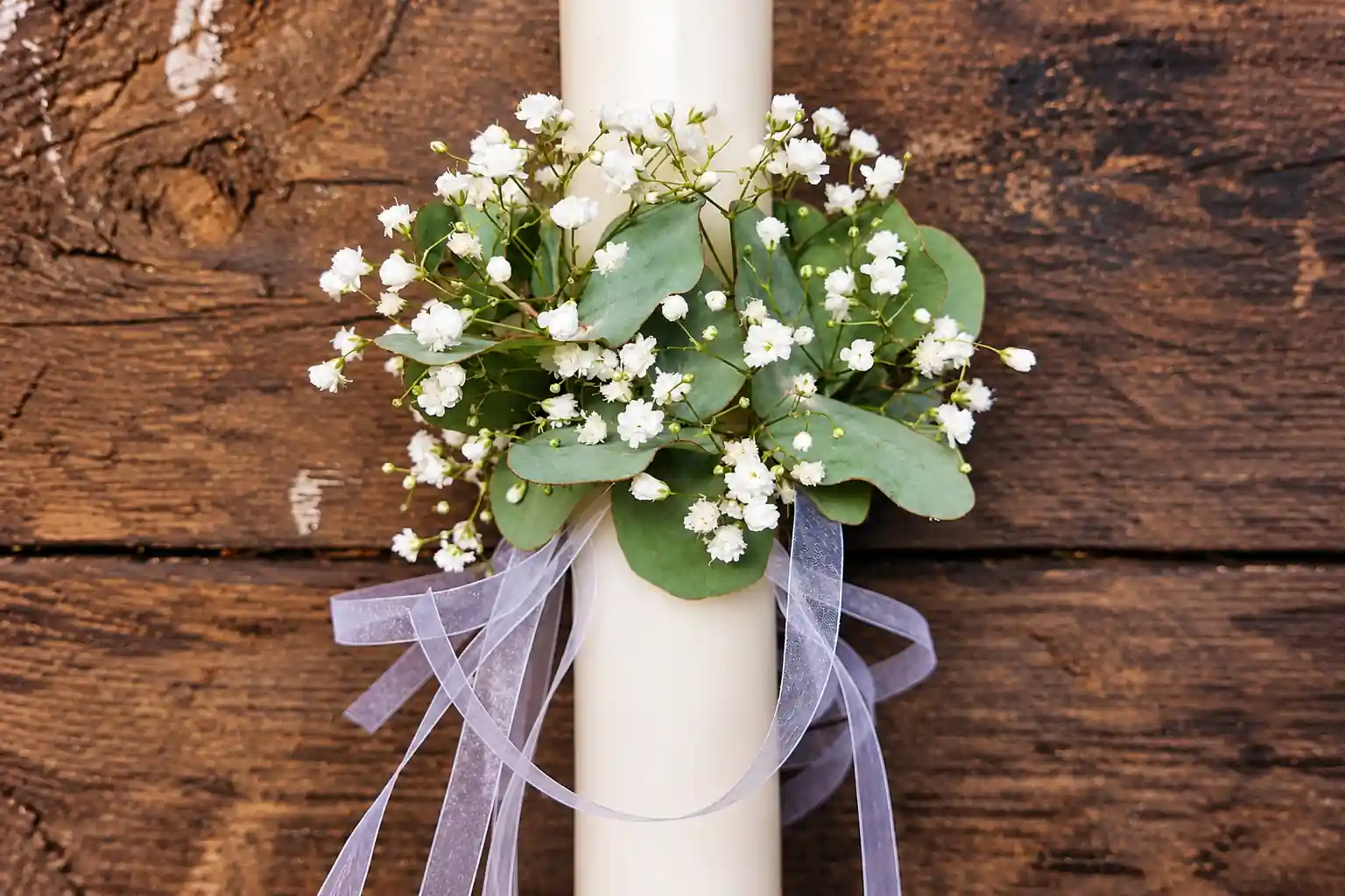 Bougie de baptême décorée de gypsophile blanc, de feuillage vert et de rubans transparents.